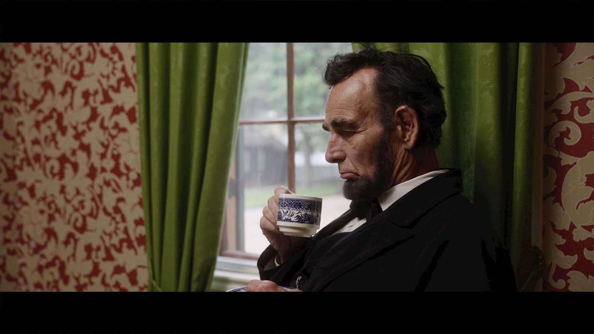 Lincoln drinks a from a cup while sitting by a window with green curtians.